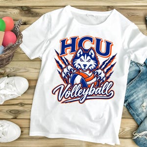 May include: A white short-sleeve t-shirt featuring a graphic of a husky mascot with blue and orange accents. The text HCU is printed above the mascot, and the word Volleyball is printed below in a stylized blue and orange font.