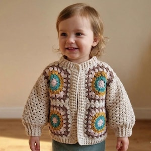 May include: A beige crochet cardigan with a button-up front. The sweater features square floral patterns in brown, teal, yellow, and pink. The sleeves are long and puffy. This handmade garment is suitable for children.