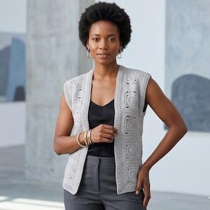 Puede incluir: Un chaleco de crochet gris claro sin mangas con un patrón cuadrado. El chaleco está abierto por delante y tiene un ribete acanalado. La modelo lleva un top negro y pantalones grises. El fondo es de color neutro.