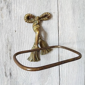 Peut inclure: Un anneau porte-serviettes en laiton doté d'une fixation murale décorative en forme de nœud de corde avec deux pompons pendants. Le métal présente une patine chaude et vieillie avec des taches d'oxydation sombres sur l'anneau et la fixation.