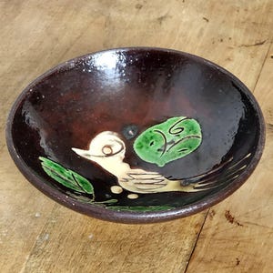 Peut inclure: Petit bol en céramique fait à la main, intérieur et extérieur brun foncé. Le bol présente un motif peint d'un oiseau et de feuilles vertes. L'oiseau est de couleur crème avec des détails noirs et jaunes. Les feuilles sont vertes.