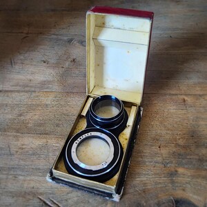 May include: Vintage camera lens set in a red and cream colored box. The lenses are black with silver trim and are labeled "SEMFLEX". The box is open, revealing the lenses nestled inside.