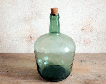 Puede incluir: Una botella de vidrio verde claro con cuerpo redondo y tapón de corcho. La botella tiene un aspecto vintage, con ligeras imperfecciones en el vidrio. La botella probablemente se utiliza para almacenar líquidos. Altura aproximada de 25 cm.