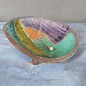 Peut inclure: Un plat ovale en céramique avec un bord brun texturé et moucheté, et un intérieur multicolore présentant des sections abstraites violettes, vertes, jaunes et orange avec des détails de lignes incisées. Un collier à chaîne dorée avec une perle blanche.
