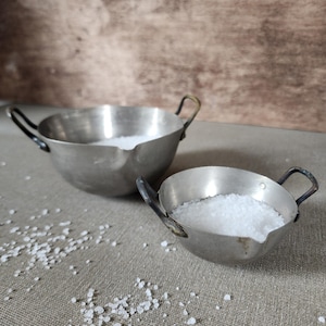 Peut inclure: Deux petits bols métalliques vintage avec des poignées, remplis de sel blanc. Les bols ont une apparence légèrement ternie, suggérant l'âge. Des cristaux de sel sont dispersés autour des bols sur une surface de ton neutre.