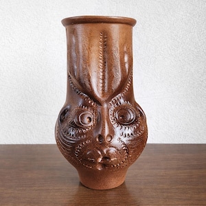 Peut inclure: Un vase en céramique marron avec un motif de visage détaillé. Le vase a une forme cylindrique avec une large ouverture et est décoré de motifs sculptés et de traits du visage, notamment des yeux, un nez et une bouche. Le vase est sur une surface en bois.