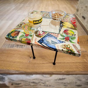 Può includere: Un tavolino quadrato con sottili gambe in metallo nero e un piano d'appoggio caratterizzato da un collage di etichette di frutta e bevande in stile vintage. Un bicchiere di liquido giallo con la scritta RICARD e una ciotolina di noci sono appoggiati sopra.