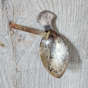 Puede incluir: Una cuchara de plata deslustrada con un mango decorativo que presenta las iniciales grabadas WM. La cuchara está atravesada por un clavo de hierro largo y oxidado que se extiende horizontalmente sobre el cuenco, creando un contraste de texturas.
