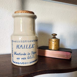 Puede incluir: Un tarro de gres vintage con tapón de corcho, etiquetado "MAILLE Moutarde de Dijon au vin blanc" en escritura azul. Una pequeña pesa de latón descansa sobre un pequeño libro. El tarro es beige con adornos azules.