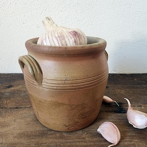 Peut inclure: Un pot à ail en céramique marron avec deux poignées et des lignes gravées. Un bulbe d'ail entier avec des stries violettes est à l'intérieur. Plusieurs gousses d'ail sont éparpillées sur la surface en bois.
