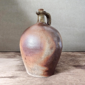 Peut inclure: Une cruche en grès antique avec un bouchon en liège. La cruche a un corps arrondi et une anse. La palette de couleurs comprend des nuances de marron, de beige et de violet, avec une finition brillante. La cruche est probablement utilisée pour stocker des liquides.