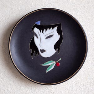 Peut inclure: Assiette en céramique noire avec un motif de visage stylisé en blanc et noir. L'assiette présente un visage de femme avec des cheveux noirs et un accent bleu. En dessous, une branche avec des feuilles vertes et une baie rouge. Bord marron.