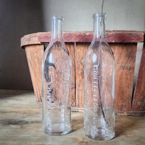 Peut inclure: Deux bouteilles en verre transparent avec des graduations, probablement pour des médicaments ou des liquides. Les bouteilles ont un aspect vintage, avec du texte en relief et des indicateurs de volume gradués. Un panier en bois est en arrière-plan.