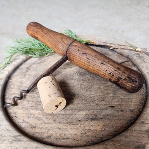 Op de afbeelding: Een vintage houten kurkentrekker met een metalen spiraal en een kurk. De kurkentrekker heeft een donkerhouten handvat en ligt op een houten oppervlak. De kurk is cilindrisch en lichtbruin.