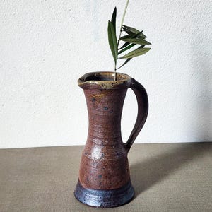 Peut inclure: Pichet en céramique marron fait à la main avec une anse, contenant une branche de feuilles vertes. Le pichet a une base bleu foncé et une surface texturée. Le bord est de couleur marron clair. Un vase rustique unique pour la décoration intérieure.