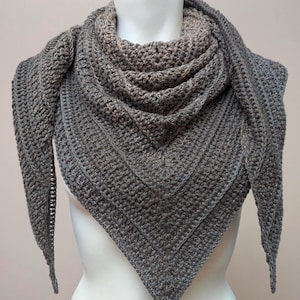 May include: A grey crochet shawl, displayed on a mannequin. The shawl has a triangular shape and a textured stitch pattern. The colour is a mix of grey and brown, creating a heathered effect. The shawl is designed to be worn around the neck and shoulders.