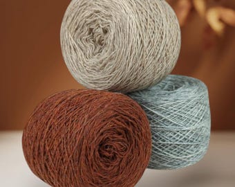 May include: Three round balls of yarn stacked together. The top ball is a light beige, the bottom left ball is a deep rust brown, and the bottom right ball is a soft sage green. Each ball features a textured, fibrous appearance with visible strands.
