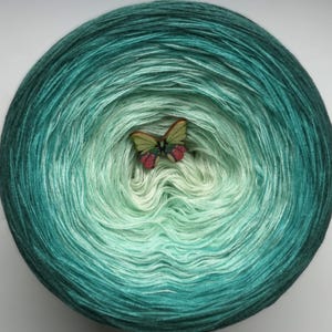 May include: A round skein of yarn in shades of teal and mint green, with a wooden butterfly embellishment in the center. The yarn's color transitions from dark teal to light mint. The butterfly has green wings and red accents.