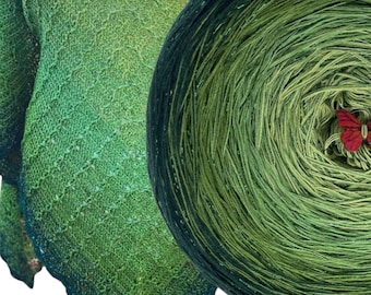 May include: A hand-knitted shawl featuring a gradient of green shades from dark to light, showcasing an intricate openwork diamond pattern. Beside it sits a circular yarn cake in matching green tones, adorned with a small red butterfly charm.