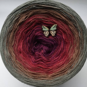 May include: A ball of yarn with a gradient of green, brown, red, and purple colours. A small wooden butterfly is placed on top of the yarn ball.