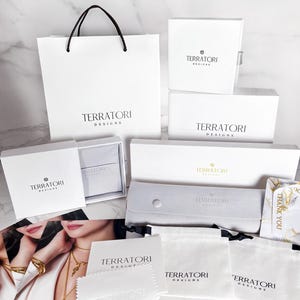 May include: A collection of white gift boxes and bags with the logo "Terratori Designs" printed on them. The boxes and bags are arranged on a white marble surface. There are also several white cloth bags with the same logo. One of the bags has a "Thank You" tag attached to it.