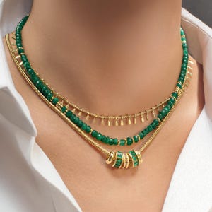 May include: A layered gold necklace with green gemstone beads and a charm. The charm is made of gold and has a ring of green gemstones.