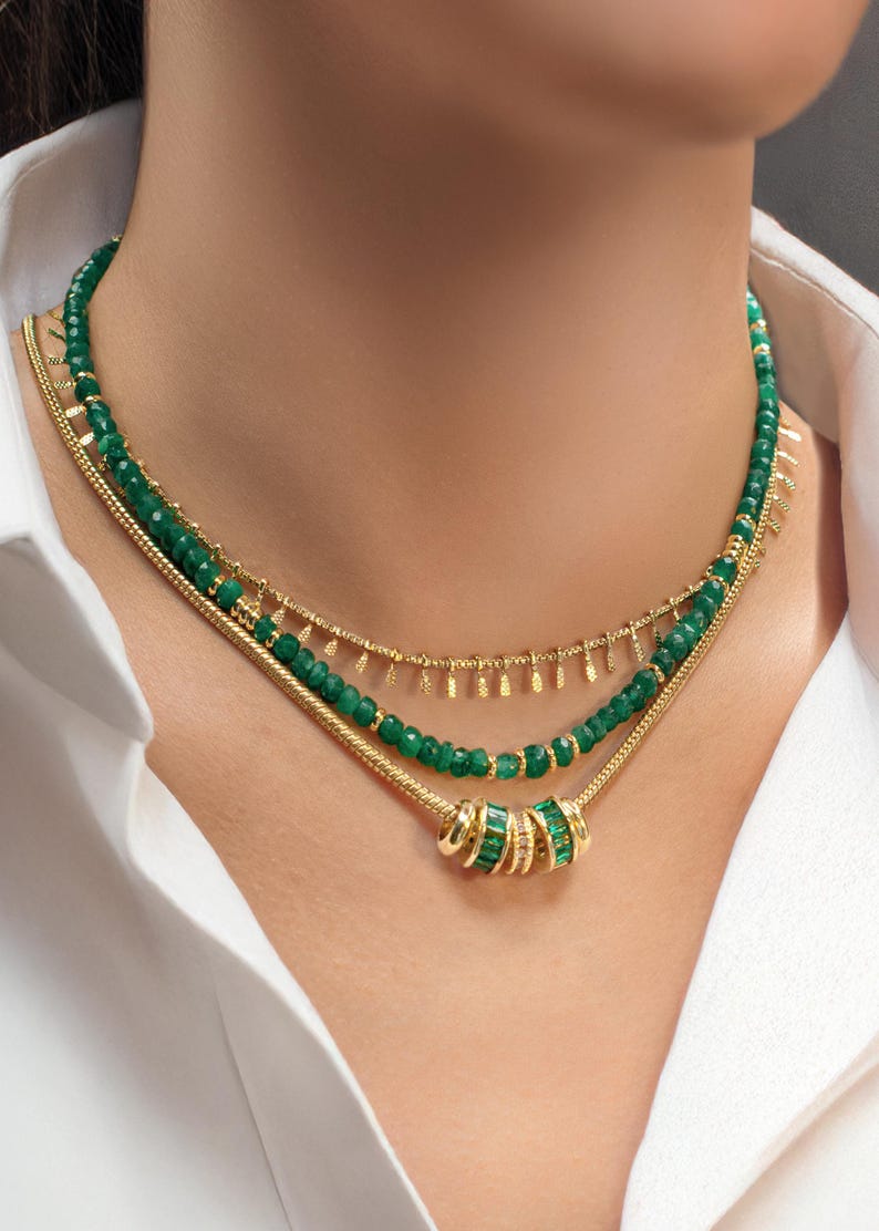 May include: A layered gold necklace with green gemstone beads and a charm. The charm is made of gold and has a ring of green gemstones.