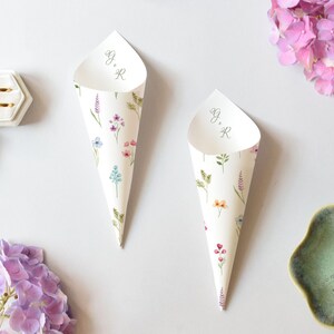 Può includere: Due coni di carta bianca decorati con fiori ad acquerello e le iniziali "G & R". I coni servono per contenere coriandoli o petali di fiori. Fedi nuziali d'oro in una scatola esagonale bianca. Fiori rosa e viola sullo sfondo.