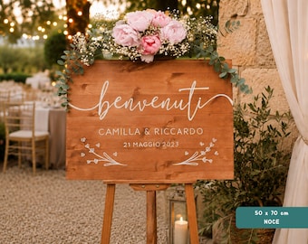Peut inclure: Un panneau en bois rectangulaire de 50 par 70 cm portant l'inscription "benvenuti", "CAMILLA & RICCARDO" et "21 MAGGIO 2023" en lettres blanches. Le panneau présente des motifs floraux blancs et est surmonté de pivoines roses et de feuillage.