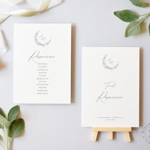 Può includere: Due biglietti da tavolo per matrimoni bianchi con calligrafia elegante. Un biglietto elenca i nomi, l'altro dice "Tavola Rosmarino" con una data. Entrambi i biglietti presentano una ghirlanda floreale e iniziali. I biglietti sono su una superficie grigio chiaro.