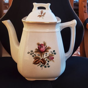 May include: A white ceramic teapot with a floral design featuring a pink rose and green leaves. The teapot has a lid with a gold trim and a floral design.