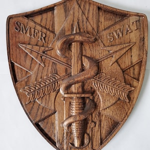 May include: A wooden shield with a carved design of a snake wrapped around a dagger, with arrows and the words "SMFR" and "SWAT" on either side.