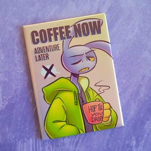 Puede incluir: Botón rectangular con un personaje de conejo de dibujos animados con una sudadera con capucha verde y sosteniendo una taza que dice "HOP TIL YOU DROP". El botón dice "COFFEE NOW / ADVENTURE LATER" con una X sobre el texto de aventura.