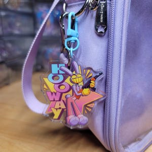 Könnte beinhalten: Ein durchsichtiger Acryl-Schlüsselanhänger mit einer Cartoon-Hasenfigur, die ein Gewehr hält, und dem Text HOOWAY in großen blauen und violetten Buchstaben. Der Anhänger ist mit einem hellblauen Kunststoffkarabiner an einer lavendelfarbenen Tasche befestigt.