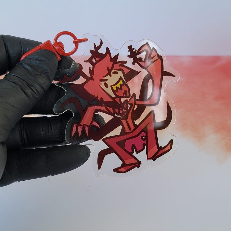 K&ouml;nnte beinhalten: Ein durchsichtiger Acryl-Schl&uuml;sselanh&auml;nger mit einer roten und schwarzen Comicfigur mit H&ouml;rnern und einer Fliege. Der Schl&uuml;sselanh&auml;nger hat einen roten Metallring.