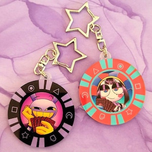 May include: Two round acrylic keychains with cartoon characters holding playing cards. One keychain is black and purple, the other is red and teal. Both have star-shaped metal charms attached.