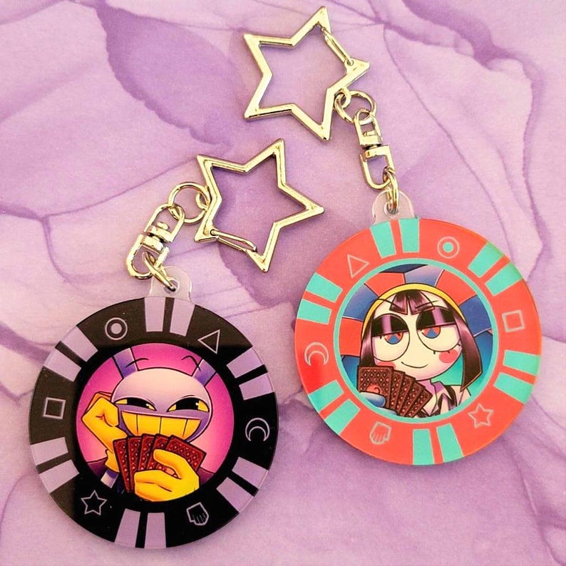 May include: Two round acrylic keychains with cartoon characters holding playing cards. One keychain is black and purple, the other is red and teal. Both have star-shaped metal charms attached.