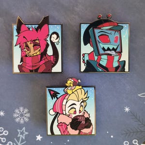 Puede incluir: Tres pines de esmalte con personajes de dibujos animados con ropa de invierno. Los pines tienen un borde cuadrado con un fondo nevado. Uno presenta un personaje con pelo rosa y una bufanda, otro tiene una cabeza con forma de televisor y el tercero sostiene una taza que dice "DEER AS".