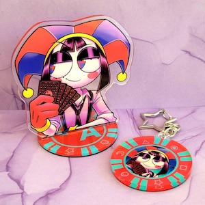 May include: A clear acrylic stand-up figure and a keychain featuring a cartoon character in a jester hat holding playing cards. The stand has a red base with the letter "A". The keychain is a red circle with a star-shaped clasp.