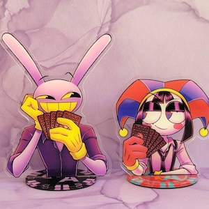 May include: Two acrylic standees featuring cartoon characters. One is a pink rabbit with yellow gloves and a purple suit, holding cards. The other is a jester-like character with a red, blue, and yellow hat, also holding cards. Both standees have detailed bases.