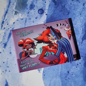 May include: A cartoon magnet featuring two characters from the show "Hazbin Hotel". The characters are a red demon with horns and a red-haired woman with a black dress. The text on the magnet reads "Are you serious?? Right in front of my voot floops!?" and "Hot."