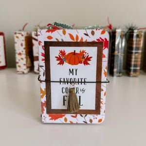 May include: A small rectangular notebook with a white cover featuring an orange pumpkin and autumn leaf pattern. The center has a brown frame with the text "MY FAVORITE COLOR IS FALL" and a small beige tassel attached to a thin black elastic band.