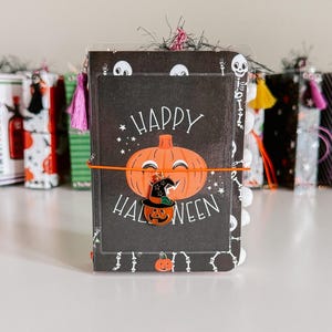 May include: A small notebook with a black cover featuring a white skeleton pattern and a central orange pumpkin illustration with the text Happy Halloween. An orange elastic band holds a small pumpkin charm, and a pink tassel hangs from the top.