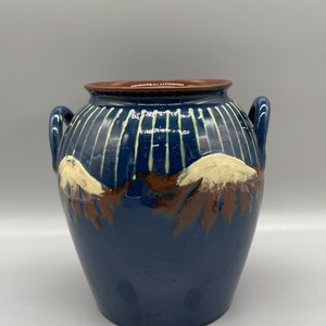 Può includere: Un vaso in ceramica blu con disegni dipinti in bianco e marrone. Il vaso ha due manici e un corpo arrotondato.