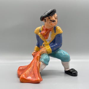 May include: A ceramic figurine of a bullfighter in a blue and yellow costume, holding a red cape. The figurine is in a crouching position, with one leg bent and the other extended.