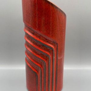 Könnte beinhalten: Eine rote Keramikvase mit geometrischem Design. Die Vase hat eine strukturierte Oberfläche und eine leicht abgewinkelte Oberseite.