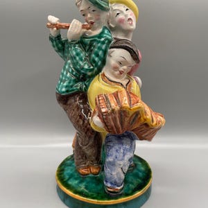 Könnte beinhalten: Eine Keramikfigur von drei Musikern. Der Musiker links trägt ein grün-weiß kariertes Hemd und einen grünen Hut und spielt Flöte. Der Musiker in der Mitte trägt ein rosa Hemd und einen gelben Hut. Der Musiker rechts trägt eine blaue Jeans und ein gelbes Hemd und spielt Akkordeon.