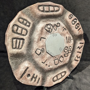 May include: A circular, weathered, brown and grey stone-like object with etched symbols and a silver, octagonal centre. The object is propped up on a black stand, suggesting it is a decorative piece or a prop.