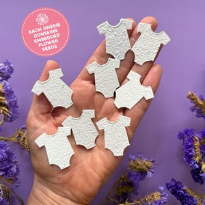 May include: A hand displays several small, white paper onesie shapes. The paper has a textured finish. A pink circle with text that reads "EACH ONESIE CONTAINS EMBEDDED FLOWER SEEDS" is in the upper left. Purple flowers are in the background.