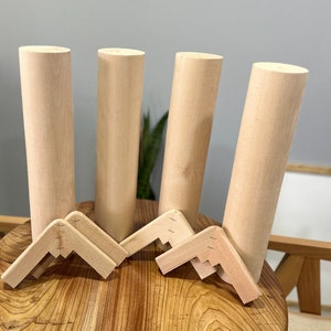 May include: Four unfinished wooden table legs and four corner brackets. The legs are cylindrical and light-colored. The brackets are angled and designed to connect the legs to a tabletop. The items are displayed on a wooden table.